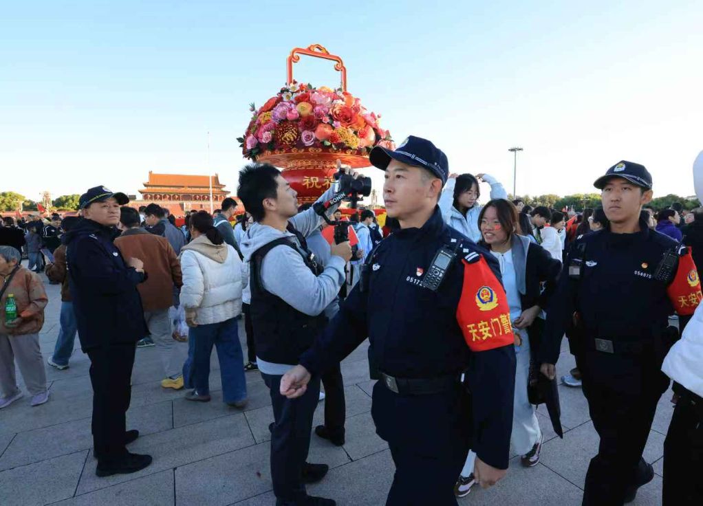 天安门广场民警在人群中执勤,背景可见国庆花篮。