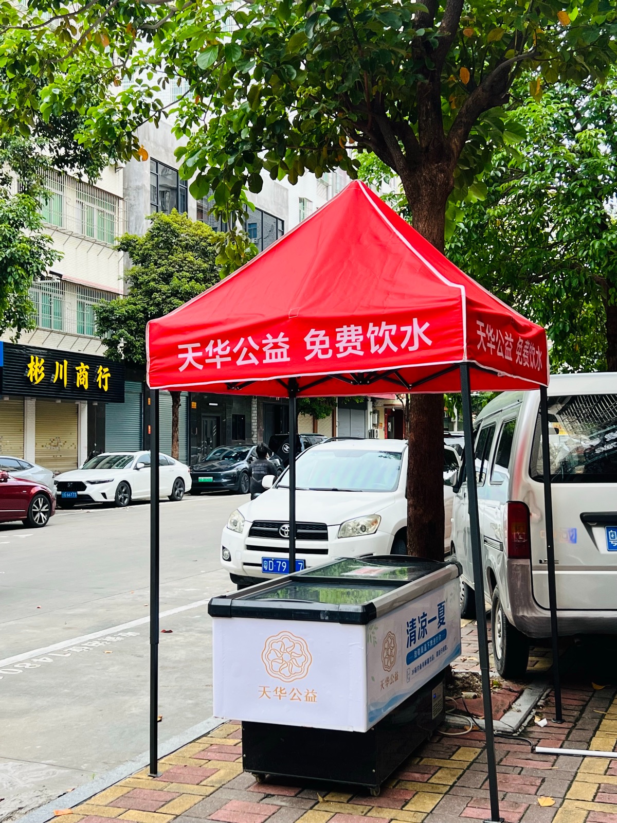 树荫下设有红色天华公益免费饮水帐篷和冷藏柜